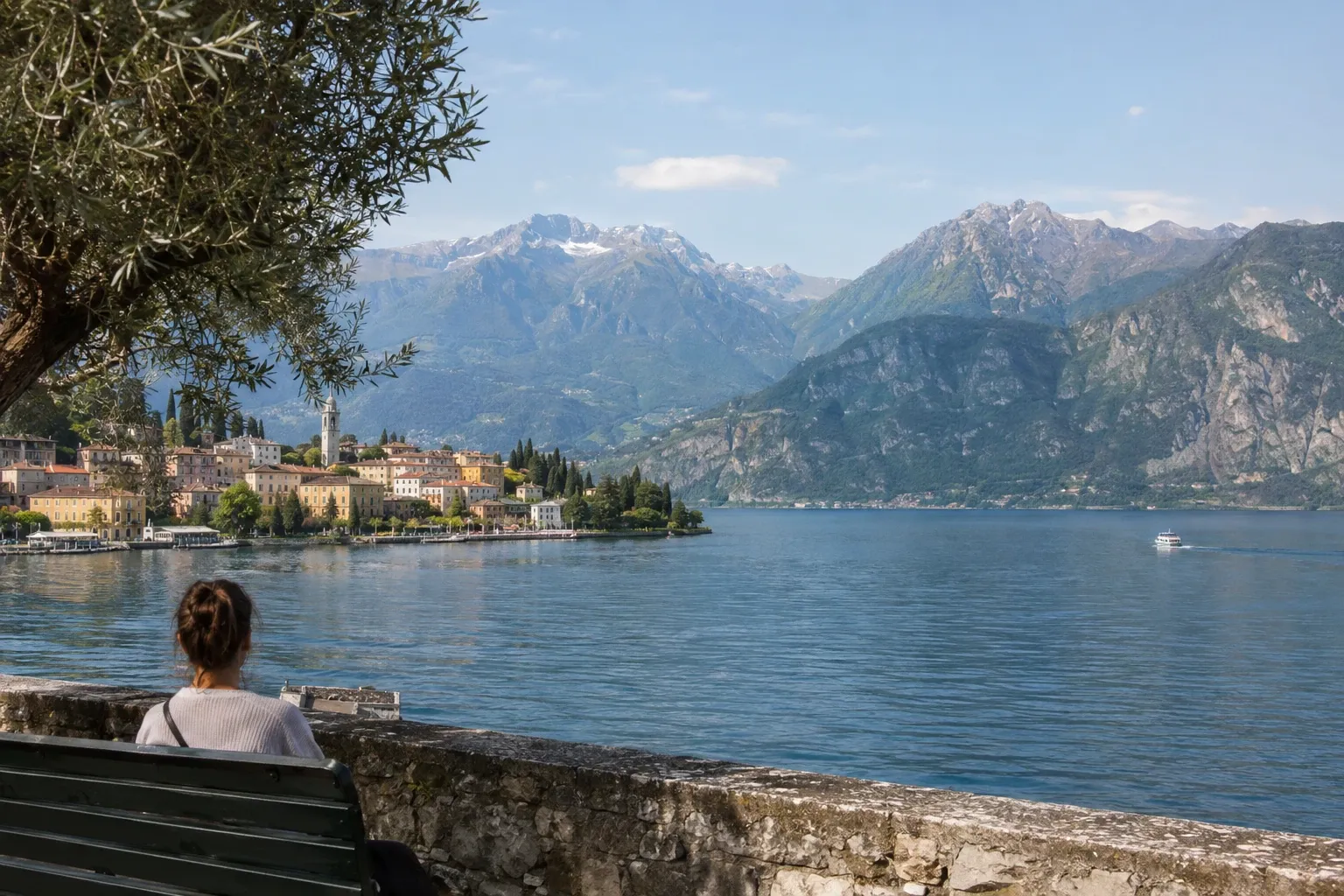 Donna seduta su panchina che guarda il lago, borgo e montagne sullo sfondo